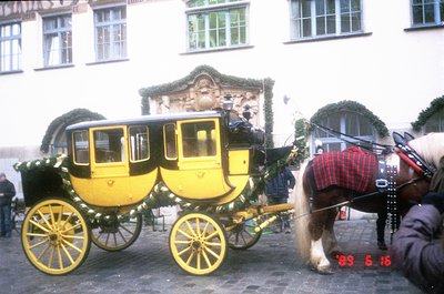 Classic yellow horse-drawn carriage with ornate brass detailing, pulled by two horses in harness. Cobblestone street flanked ...