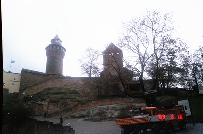 Medieval stone fortress with two cylindrical towers on elevated rocky terrain. Overgrown vegetation and a vintage truck (lice...