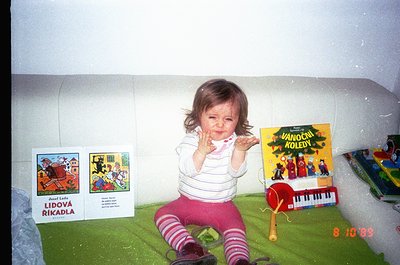A toddler sits on a green play mat in a 1990s indoor play area, clapping hands. Surrounding them are Czech children’s books: ...