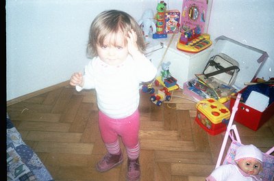 Young child in 1980s/90s indoor playroom, holding toy steering wheel. Surrounding toys include a toy piano, colorful plastic ...