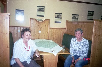 Two women seated at a wooden café table in a rustic wooden-paneled room, dated 1985. The woman on the left wears a white blou...