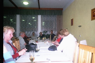 Vintage indoor gathering at a long dining table with 8 adults seated, dressed in 1980s-90s casual attire. Glassware, wine bot...