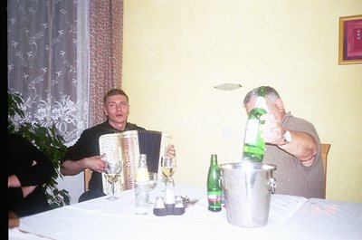Intimate gathering featuring two men at a table: one playing an accordion, the other pouring beer from a bucket. Table set wi...