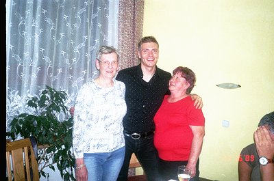 Three adults pose indoors, likely a private home or gathering space. The man in the center wears a black patterned shirt and ...