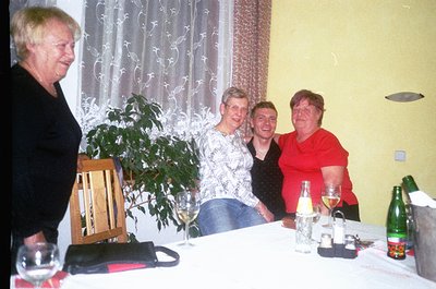 Four women gather indoors at a table set with wine glasses, a bottle, and floral decor. The setting suggests a casual, mid-20...