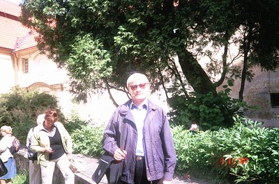 Two adults in 1970s-style attire pose outdoors in a lush, shaded garden. The man wears a dark jacket, sunglasses, and a light...