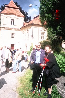 Neatly framed 1990s-era photo of elderly couple in a European courtyard. Man in light blue sweater, sunglasses, and cane; wom...