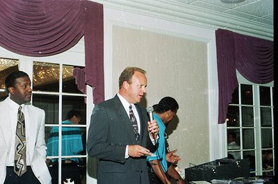 Formal indoor event with three men in 1990s business attire—dark suits, ties, and dress shirts. Central figure holds a microp...