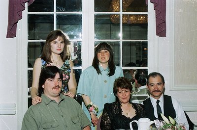 Five adults pose indoors near a large window with decorative trim, likely a 1980s-1990s wedding or formal event. Men wear dre...