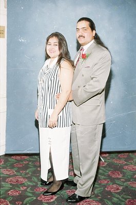 A 1990s-era formal portrait featuring a man and woman in coordinated attire. The man wears a light gray suit with a red bouto...