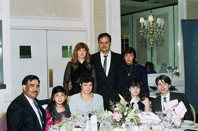 Family portrait at a formal banquet hall, likely 1980s–1990s. Seven individuals pose indoors with a chandelier and mirrored w...