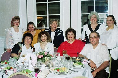 Family gathering at a formal indoor banquet, likely 1990s. Eight adults pose around a long table set with crystal glasses, fl...