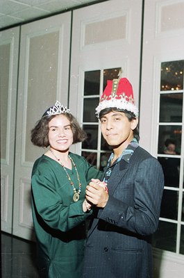 Two individuals pose indoors, likely at a festive event in the 1970s–1980s. The woman wears a tiara and green dress with a be...