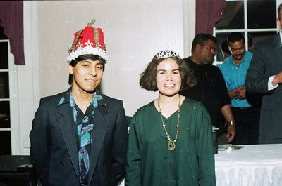 Two individuals pose indoors in festive attire, likely 1980s–1990s. The man wears a red-and-white crown, patterned blazer, an...
