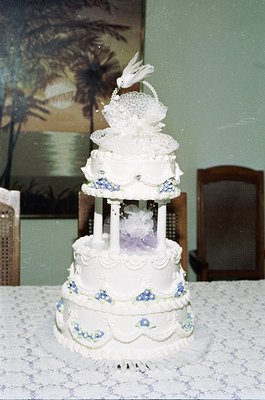 Three-tiered wedding cake with intricate white icing, floral blue accents, and a decorative bird topper on a vintage tableclo...