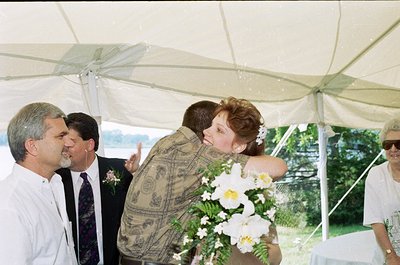 Under a white canopy tent, a bride in a floral dress and groom in a patterned shirt share an emotional moment. Surrounding th...