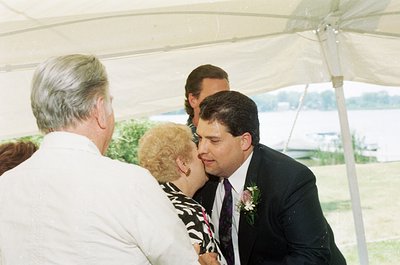 Intimate moment at an outdoor wedding ceremony under a white canopy tent. A groom in a dark suit with a pink boutonnière lean...