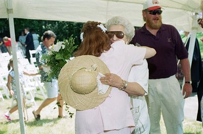Two women embrace at an outdoor event, likely a wedding or funeral. The woman in the foreground wears a wide-brimmed straw ha...