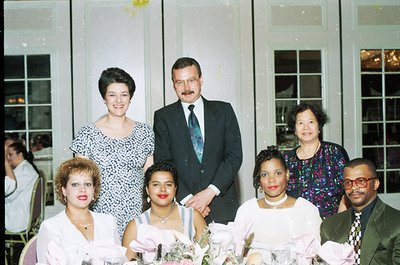 Vintage group portrait at an indoor banquet hall, likely 1980s–1990s. Six adults pose formally: three women in formal dresses...