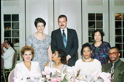 Family portrait from the **1980s–1990s**, featuring seven individuals in formal attire at an indoor banquet. The seated group...