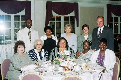 Elegant 1990s formal gathering at a banquet hall, featuring nine adults seated at a long table adorned with floral centerpiec...