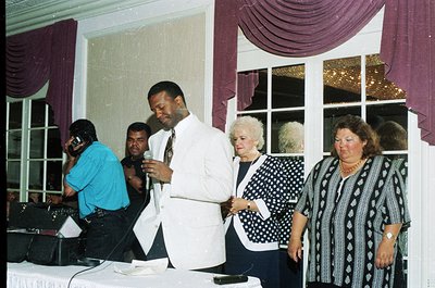 Elegant 1990s indoor event with polished wooden floors and draped maroon/purple curtains. Central figure in white suit speaki...