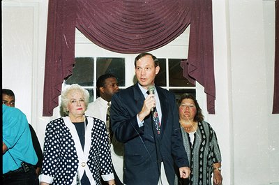 Indoor formal event featuring a man in a dark suit speaking into a microphone, flanked by three women in formal attire. The b...