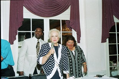 A woman in a 1990s-style polka-dot blouse holds a microphone on stage, flanked by two women in patterned blazers. Purple drap...