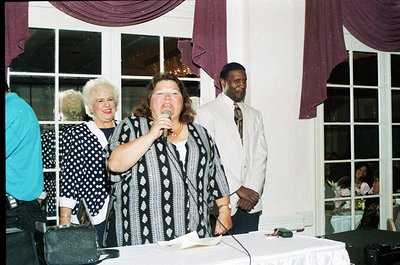A woman in a patterned blouse speaks into a microphone at a formal indoor event, likely a 1990s banquet or ceremony. Purple d...