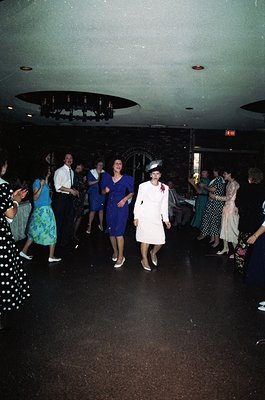 Vintage 1970s indoor dance party with retro attire—men in dress shirts, women in bold floral prints and fitted dresses. Centr...