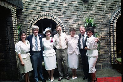 Six individuals pose in formal attire outside a brick building with an arched doorway, likely a 1970s wedding. Four women in ...