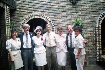 Six individuals pose outdoors in formal attire, likely a wedding or celebratory event, framed by a brick archway. The woman i...