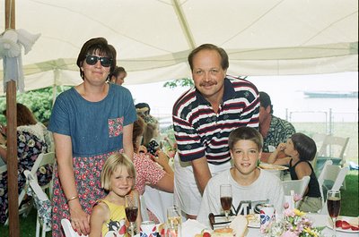 Family gathering at an outdoor event under a white canopy tent, likely a wedding or reception. Adults and children pose in 19...