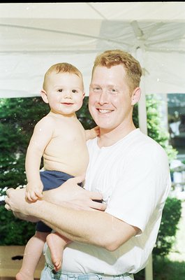 A man in a white shirt and denim shorts holds a toddler outdoors under a canopy. The child, shirtless in dark shorts, smiles ...