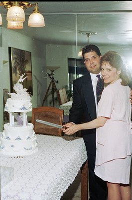 A 1970s-style wedding cake cutting scene in a vintage home. The multi-tiered cake features blue floral designs and a bird mot...
