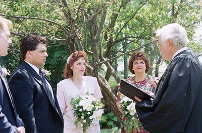 Outdoor wedding ceremony with bride in vintage 1990s attire holding white bouquet. Officiant in black robe conducts ceremony ...