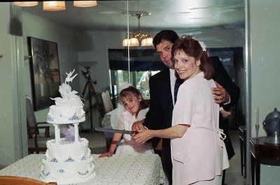 1970s-style wedding cake cutting with floral accents. Couple in formal attire (man in suit, woman in bridal gown) pose with a...