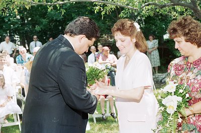 1970s outdoor wedding ceremony with bride and groom exchanging bouquets. The groom in a dark suit and the bride in a white go...