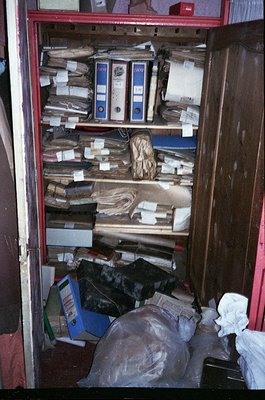 Overcrowded metal filing cabinet filled with aged binders, loose documents, and miscellaneous paper stacks. Evidence of decad...