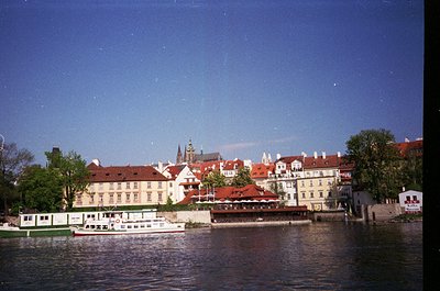 Historic European riverside scene with classic red-tiled rooftops and a prominent church spire. Mid-20th century architecture...