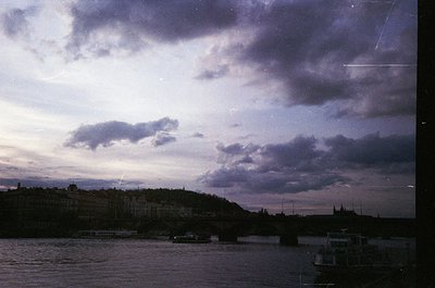 Vintage urban riverscape at dusk, featuring a bridge spanning a calm waterway. Dense, low-rise buildings line the hillside, s...