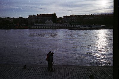Two figures stand on a cobblestone riverside promenade, silhouetted against a vintage riverboat and urban skyline. The boat’s...