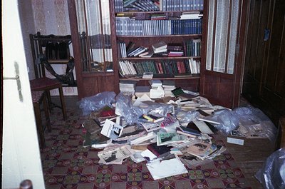 Vintage room in disarray, featuring wooden paneling and a tiled floor with floral patterns. Shelves overflow with books, mixe...
