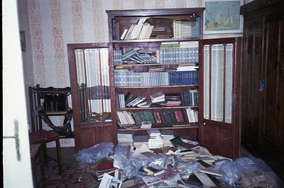 Vintage wooden bookcase filled with books, likely from the 1970s–1990s, in a room with faded floral wallpaper. Floor littered...