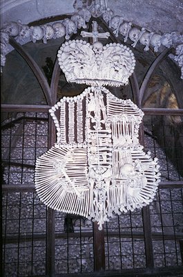 Memento mori ossuary art featuring skulls and bones arranged into a crown, cross, and floral motifs atop a stone arch. Likely...