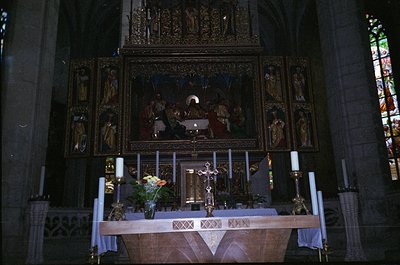 Gothic-style altar with intricate wooden carvings, flanked by candle holders and floral arrangements. Central mural depicts r...