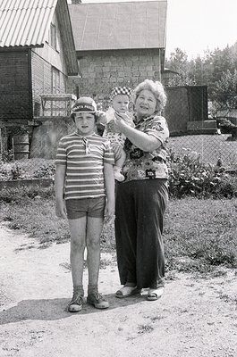 A black-and-white snapshot from the 1960s–70s featuring a grandmother, child, and baby outdoors in a rural setting. The woman...