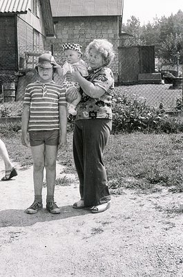 A mid-20th-century black-and-white snapshot captures a grandmother, child, and toddler in a residential backyard. The grandmo...
