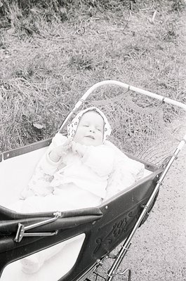 Vintage black-and-white photo of an infant in a classic metal-framed pram with plush lining, seated outdoors on a grassy path...