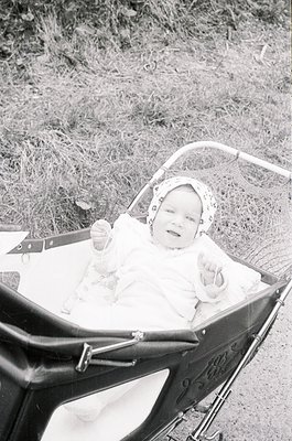 Vintage black-and-white photo of an infant seated in a classic metal-framed pram with ornate detailing, wrapped in a white fu...